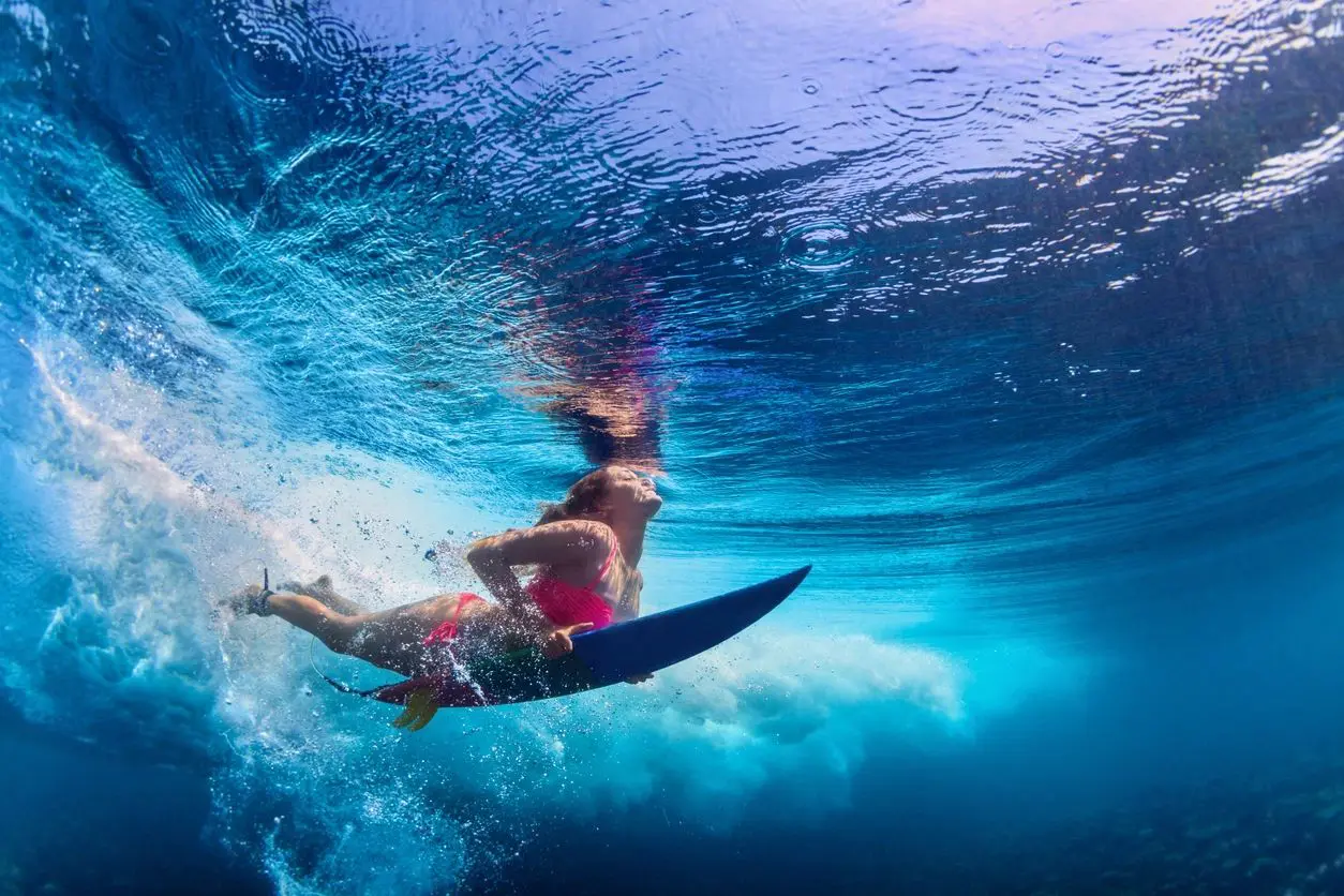 Young active girl wearing bikini in action - surfer with surf board dive underwater under big ocean wave.